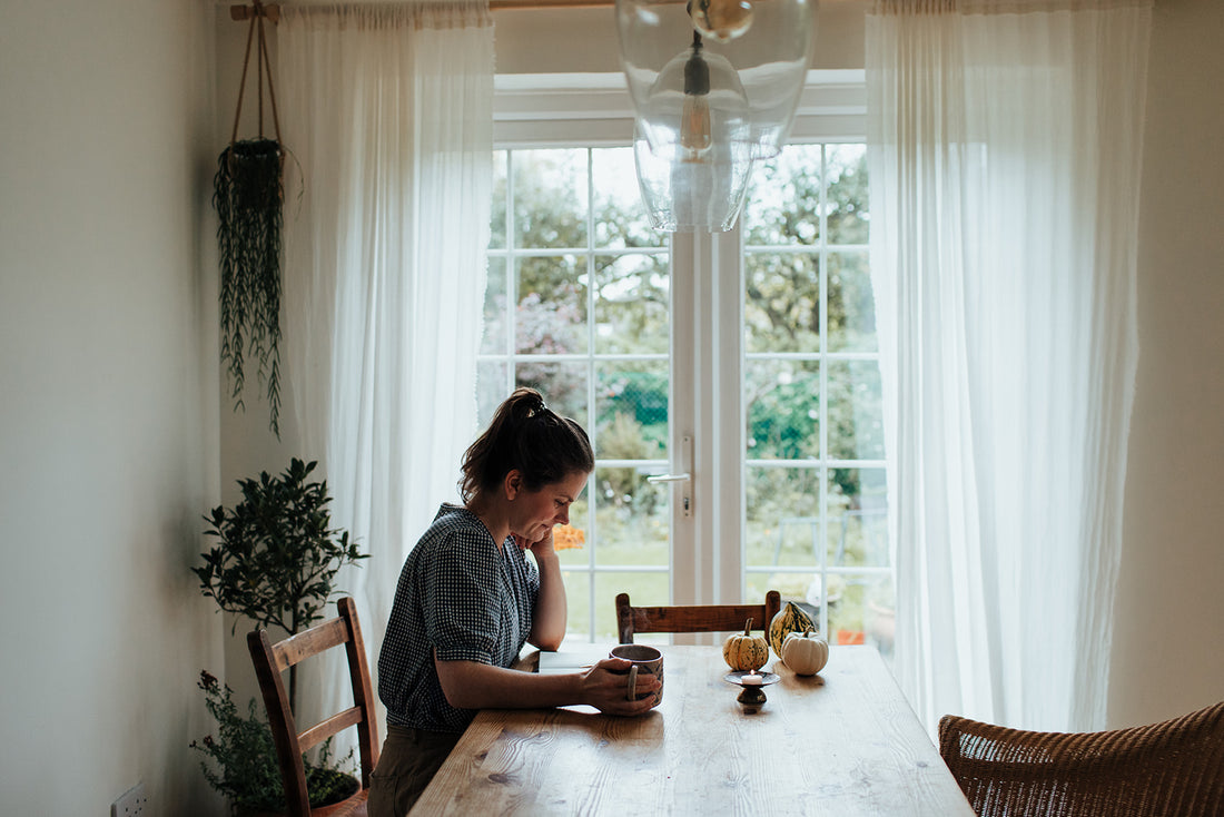 Agnes, a white woman with brown hair in her late 30s, sits at a wooden table with french doors outside behind her. She holds a cup of tea and is looking down at a book. Pumpkins are on the table.